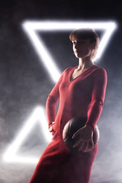 漂亮的女孩穿着漂亮的红色连衣裙 拿着篮球 带有白色霓虹灯的雾背景下的工作室肖像 — 图库照片