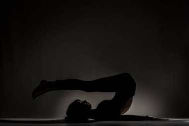 Fit balerin kız yalan söylüyor ve yoga pozları veriyor. Koyu arkaplana karşı yan aydınlatma silueti.
