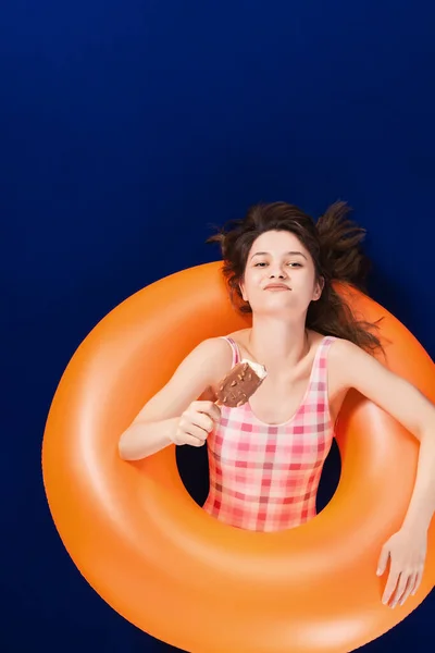 膨脹可能なオレンジ色のゴムリングに横たわっているワンピースピンクの水着で幸せな女の子とアイスクリームを楽しんでいます 夏休みのコンセプト 青い表面と背景 — ストック写真