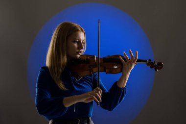 Beautiful blond girl playing violin. Female violinist against dark blue background with smoke.