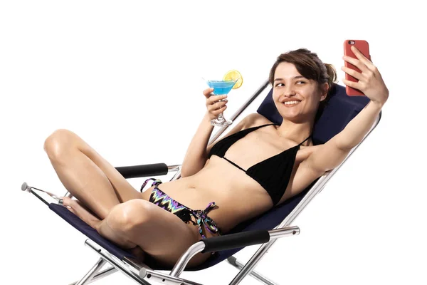 身着泳衣的女孩坐在躺椅上休息 鸡尾酒在白色背景下拍摄自拍照片 海滩概念 — 图库照片