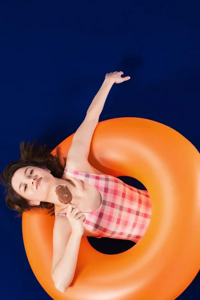 快乐的女孩穿着一件粉红色的泳衣躺在充气橙色的橡胶圈上 享受着冰淇淋 暑假的概念 蓝色表面和背景 — 图库照片