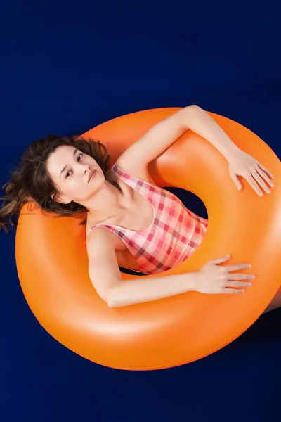 快乐的女孩穿着一件粉红色的泳衣 躺在充气橙色的橡胶圈上 暑假的概念 蓝色表面和背景 — 图库照片