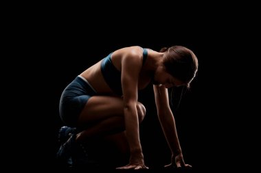 Düşük yarış başlangıç pozisyonunda kadın koşucu silueti. Siyah arka plana karşı spor giyimli bir kız.