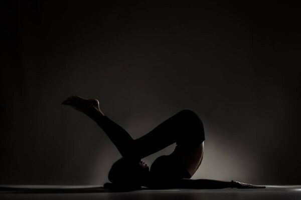 Fit ballerina girl lying and stretching practicing yoga poses. Side lit silhouette against dark background.