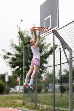 Basketbol oynayan genç bir kız. Çemberde asılı duruyor. Kentsel geçmişe sahip açık hava sporları sahası.