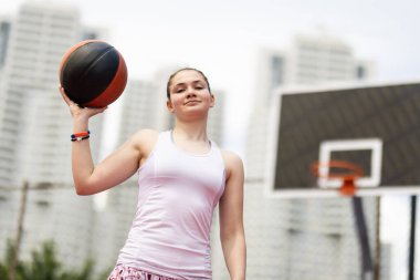 Basketbolcu portresi. Elinde topla poz veren bir kız. Kentsel geçmişe sahip açık hava sporları sahası.