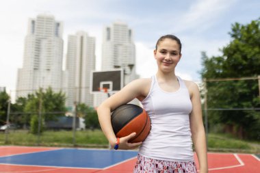Basketbol oynayan güzel bir genç kızın portresi. Topu elinde tutuyor. Kentsel çevrede açık hava spor sahası.