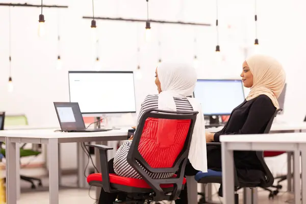 Two african american muslim girl colleagues with hijab posing together in a modern office. Monitors and light bulbs in the background and foreground
