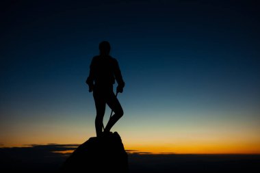 Kaya kaidesinde elleri havada gezen bir kızın silueti. Mavi gökyüzünde güzel turuncu gün batımı.