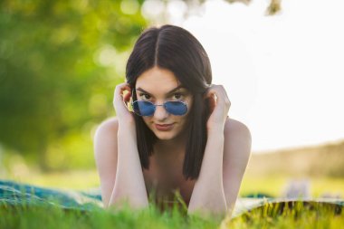 Parktaki çayırda uzanmış güneş gözlüklü güzel siyah saçlı kız. Kameraya bakıyorum..