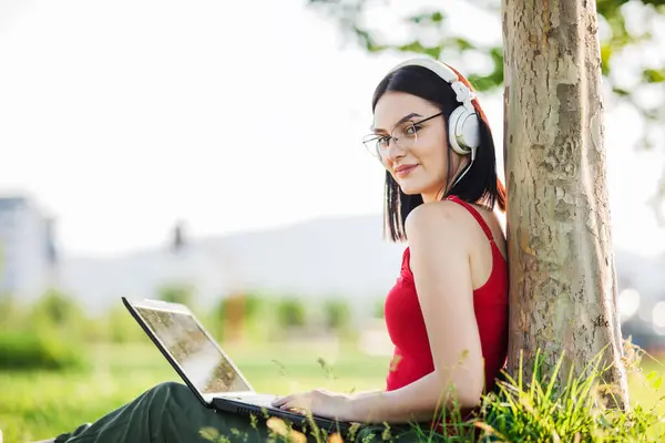 girl with dark brown hair wearing white headphones using laptop in a park under a tree