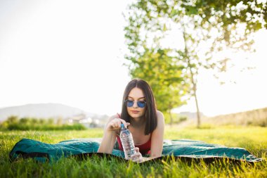 Parkta plastik bir su şişesiyle yatan güzel bir kız. Güneşli bir yaz gününde açık hava aktivitesi.
