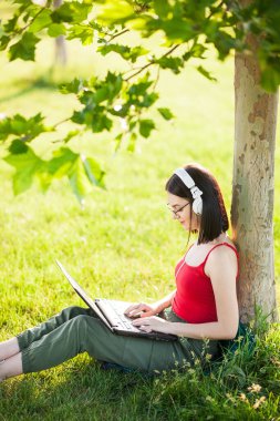 Koyu kahverengi saçlı, beyaz kulaklık takan, ağaçların altında dizüstü bilgisayar kullanan bir kız.