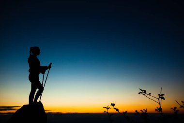 Kaya kaidesinde elleri havada gezen bir kızın silueti. Mavi gökyüzünde güzel turuncu gün batımı. Ön planda yaprakları olan ağaç dalları.