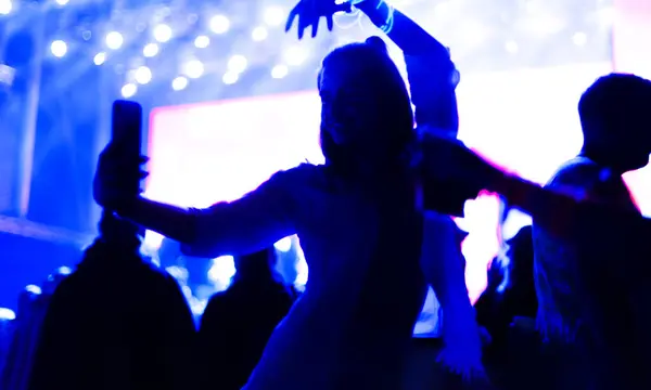 Girl stands in front of a crowd. Silhouette lady. Holding a cell phone up taking selfy photo. Out of focus festival concert stage with flashing lights in the background.