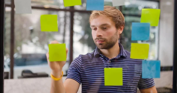 Young businessman standing near glass window with sticky notes. Man organizing startup project ideas with scrum methodology. Colorful stickers on window.