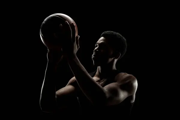 Basketball player throwing a ball against black background. Muscular african american man sidelit silhouette.