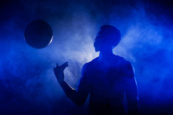 Basketball player holding a ball against blue fog background. Muscular african american man silhouette.