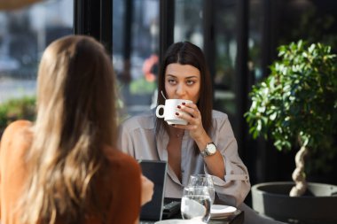 Two young business women in a cafe having one on one meeting. Friends after work talking gossiping and having coffee at a window table with reflections.