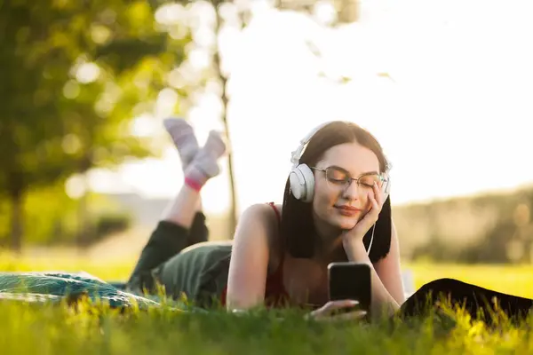 スマートフォンからヘッドフォンで音楽を聴く美しいダークヘアガール 緑色の牧草地に横たわっている ストック画像
