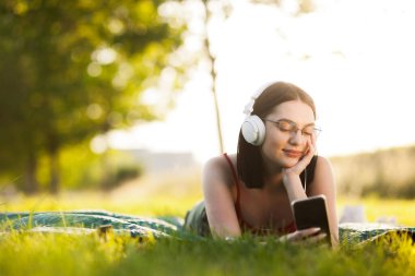 Güzel siyah saçlı kız akıllı telefonundan kulaklıkla müzik dinliyor. Yeşil bir çayırda yatıyor.