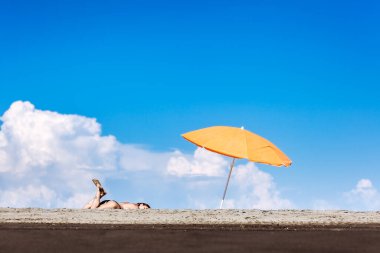 Huzurlu bir ortam, turuncu bir plaj şemsiyesinin altında uzanmış, deniz kenarında huzurlu güneşli bir günün tadını çıkaran genç bir kadını yakalıyor..