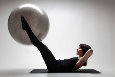 Odaklanmış genç bir kadın, hafif ışıklandırılmış bir stüdyo ortamında yoga minderinde büyük bir fitness topu kullanarak pilates seansına katılıyor..