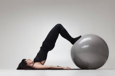 Siyah elbiseli genç bir kadın, minimalist bir stüdyo ortamında büyük bir fitness topu kullanarak güç ve denge gösterisi yapıyor..