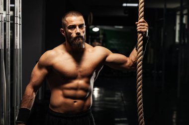 Sakallı kararlı kaslı bir adam spor salonunda tırmanma halatını tutuyor. Yoğun bir çalışma sırasında güç ve odaklanma gösteriyor..