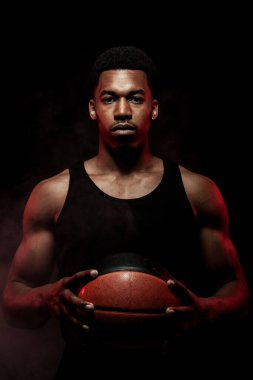 Basketbolcu tarafı kırmızı renkte, siyah arka planda topu tutuyor. Yoğunlaşmış kas yığını Afrikalı Amerikalı adam.