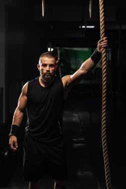 Güçlü erkek fitness meraklısı, loş ışıklı bir spor salonunda ipe tırmanmaya hazırlanıyor, kaslı ve yoğun bir odak noktası gösteriyor..