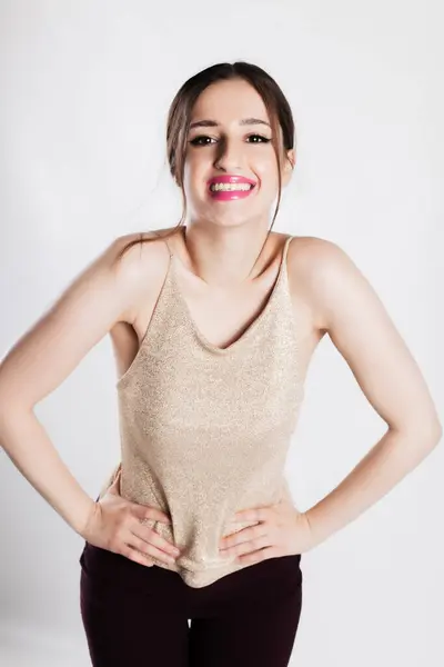 stock image A young woman stands confidently in a sparkling golden tank top, smiling brightly against a plain white background.