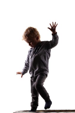 Gri kıyafetli bir çocuğun silüeti trambolinde tek eliyle dengede duruyor. Hareketi, dengeyi ve çocukluğu neşeli bir anda yakalar.