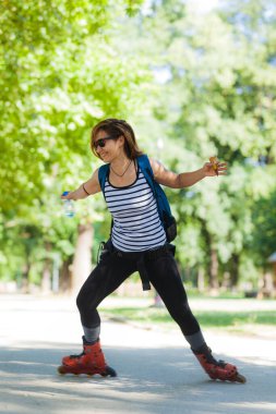 Neşeli bir kadın parkta paten kayıyor, dengeli ve gülümsüyor, ağaçlar yemyeşil bir zemin sağlıyor. Günlük yaz kıyafetleri giyiyor ve bir sırt çantası taşıyor..