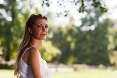 Beyaz kolsuz elbise giyen orta yaşlı bir kadın, ağaçların arasından süzülen güneş ışığıyla, yemyeşil bir parkta güven içinde duruyor..