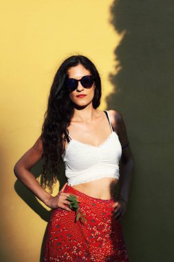 Bu resim, beyaz bir ekin bluzu, çiçekli kırmızı bir etek ve güneş gözlüğü giymiş, parlak sarı bir duvarın yanında duran kendine güveni tam genç bir kadını yakalıyor..