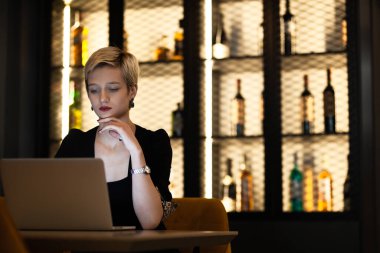 Siyah kıyafetli genç sarışın bir kadın. Arkasında güzel aydınlatılmış bir şişe rafı olan bir barda dizüstü bilgisayarına odaklanıyor..