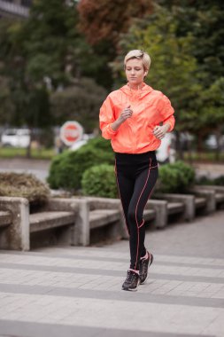Parlak turuncu ceketli ve siyah taytlı odaklanmış genç bir kadın bir şehir parkında koşuyor. Forma ve sağlıklı bir yaşam tarzına bağlılığını resmediyor..