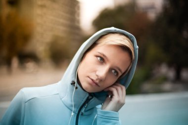 Mavi kapüşonlu genç bir kadının portresi, kulaklıkla müzik dinliyor, açık hava şehir arka planında..