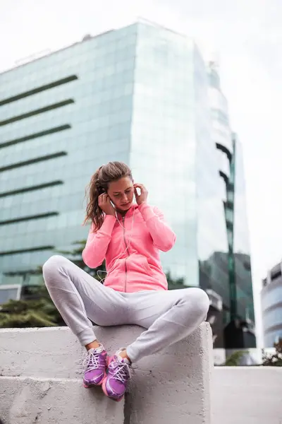 stock image Fitness-focused young female relaxes on a ledge with her headphones on, in a modern cityscape backdrop, wearing a bright pink hoodie.