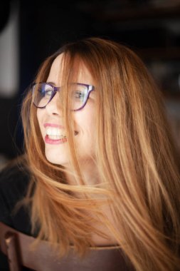 Kızıl saçlı ve gözlüklü neşeli bir kadının bulanık bir arkaplanla gülüşünü gösteren samimi bir görüntü..