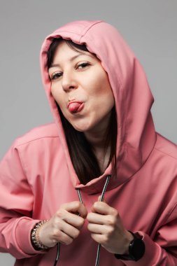 Pembe kapüşonlu, neşeli bir genç kadın, tarafsız bir arkaplana karşı arsız bir yüz ifadesiyle dilini dışarı çıkarıyor..