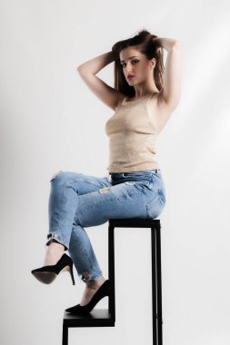 Parlak bluzlu ve yırtık pırtık kotlu şık bir bayan model, siyah bir taburede zarif bir şekilde oturuyor kendine güveni ve tarzıyla.