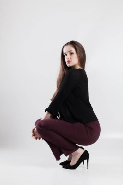 Siyah bluz ve kestane rengi pantolon giyen şık bir genç kadın omuzlarının üzerinden temiz beyaz bir zemin üzerine çömelmiş bakıyor..