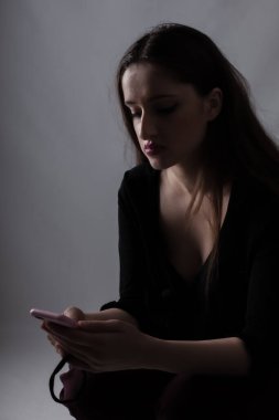 Koyu renk giyinmiş genç bir kadın telefonunu karanlık bir odada kullanıyor. İç gözlem ve yalnızlık havasını yansıtıyor. Görüntü, sessiz ve yansıtıcı bir anı tasvir ediyor..