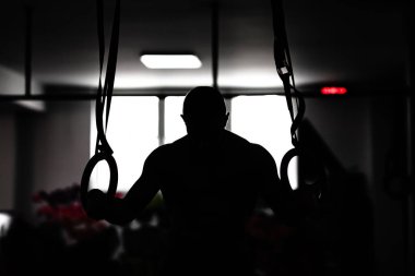 Spor salonunda elinde jimnastik yüzükler tutan güçlü bir erkek atlet silueti. Gücünü ve dikkatini gösteriyor..