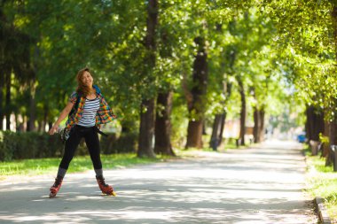 Güneşli bir günün tadını çıkaran enerjik bir kadın yapraklı bir parkta paten kayarken renkli günlük giysiler giyer ve neşeyle gülümserdi..