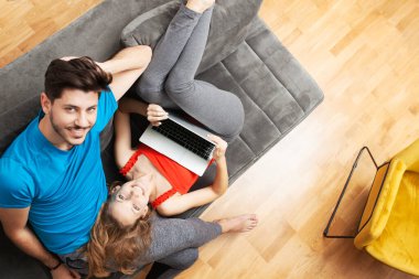 Gri bir kanepede uzanan mutlu bir çift rahat bir ev ortamında dizüstü bilgisayarla meşgul oluyor..