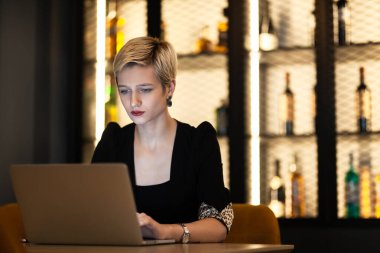 Şık siyah kıyafetli, odaklanmış genç iş kadını canlı bir ofis kafesinde dizüstü bilgisayarıyla çalışıyor. Modern çalışma tarzı ve konsantrasyonu sergiliyor..